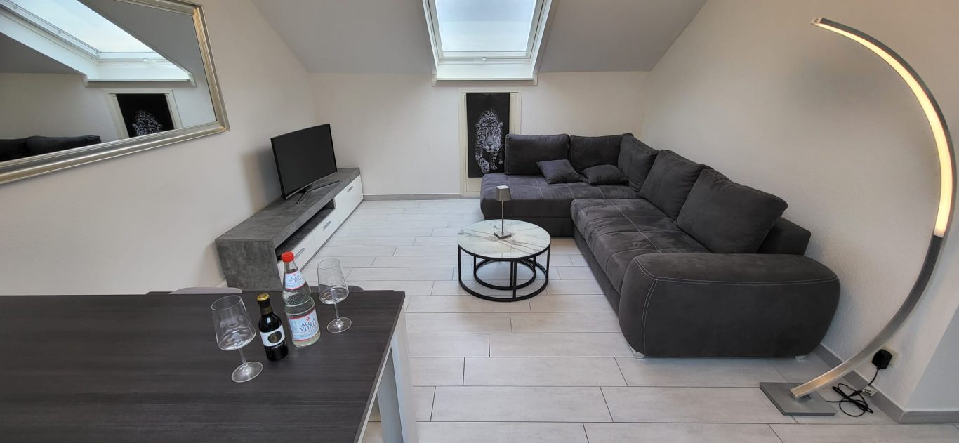 Heller Wohnraum mit grauem Sofa, Tisch und Lampe unter einem Dachfenster.